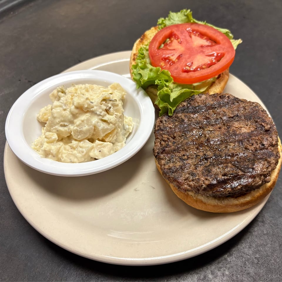 A grilled burger patty on a bun with lettuce and tomato, served with a side of potato salad.
