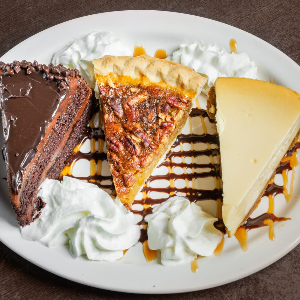 A dessert plate with slices of chocolate cake, pecan pie, and cheesecake, garnished with whipped cream and sauces.
