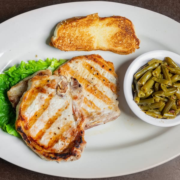 Grilled pork chop with green beans, lettuce, and toasted bread on a white plate.