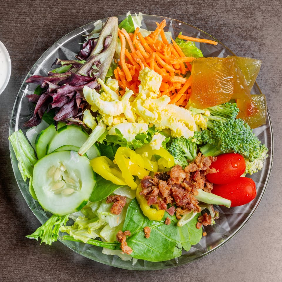 A vibrant salad with lettuce, cucumbers, tomatoes, carrots, broccoli, bacon bits, and yellow peppers.